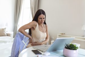 Businesswoman Holding Her Back While Working On Laptop At Home Office Desk. young woman, stretching, sitting feeling her back tired after working at laptop, small home office interior.